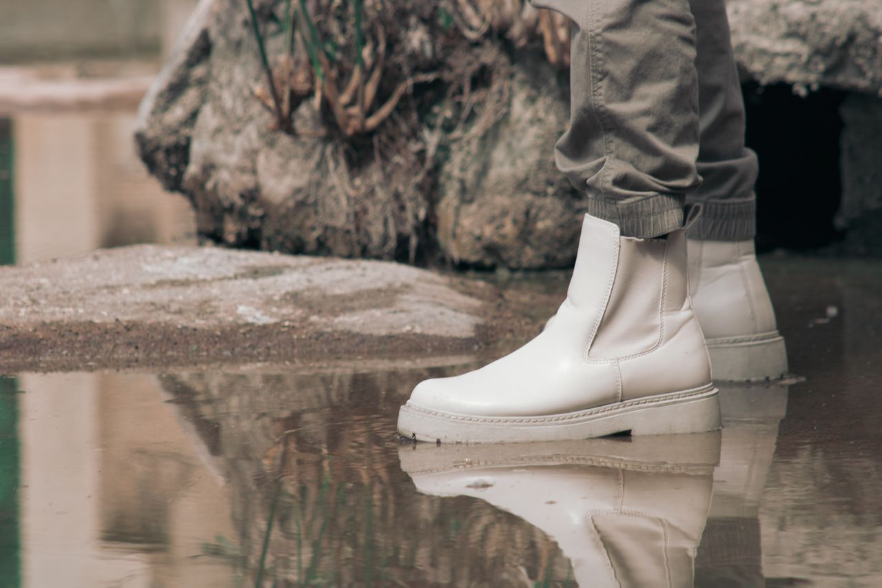 White Chelsea boots in a puddle with a rocky outdoor background, showcasing a stylish and reflective moment.
