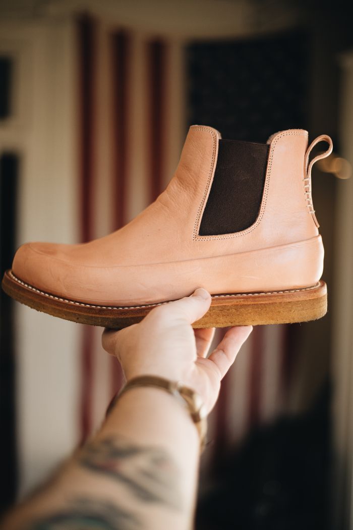 A handcrafted leather boot held in front of a backdrop featuring the American flag.