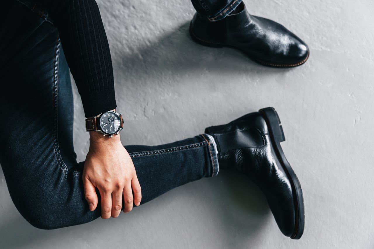 A man wearing denim jeans and leather boots, showcasing a sophisticated wristwatch.